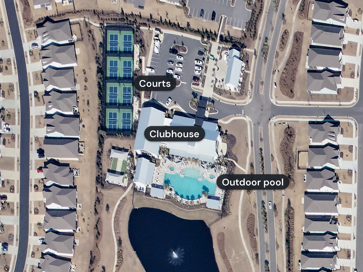 The Del Webb North Myrtle Beach clubhouse from above, labelled: a large gray-roofed building, an outdoor pool with a deck behind it, a row of green sports courts by the parking lot, and a pond and rows of homes around them