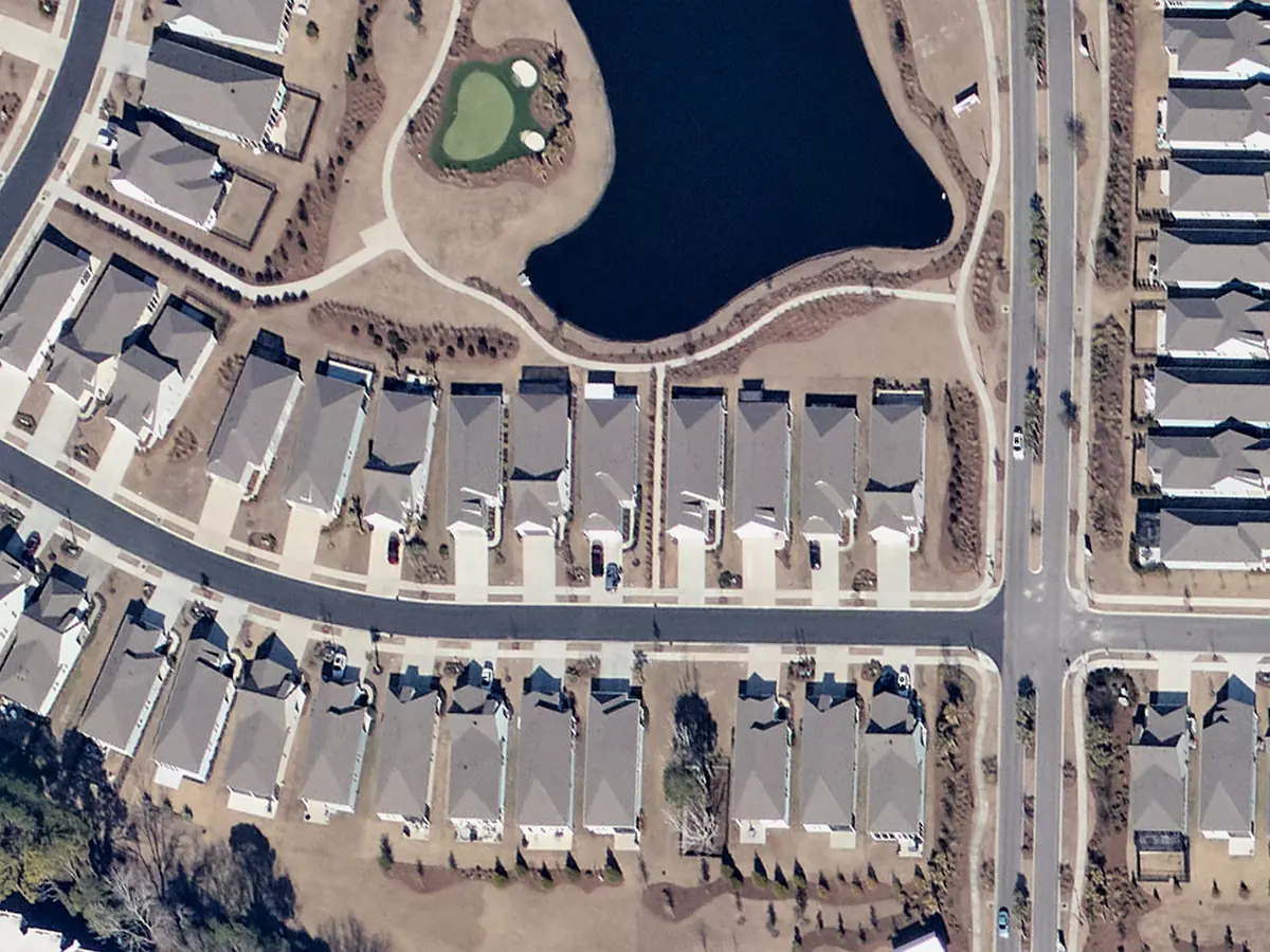 Two rows of homes with gray roofs and short driveways along a straight street in Del Webb North Myrtle Beach, with a pond and a paved walking path behind them, seen from above