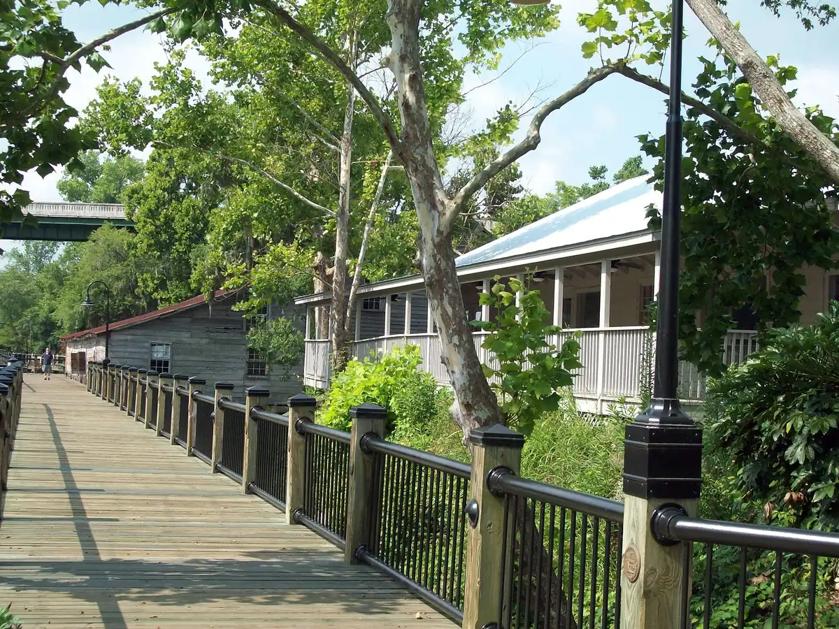A wooden boardwalk with a black railing, shaded by trees, beside old wooden buildings in Conway