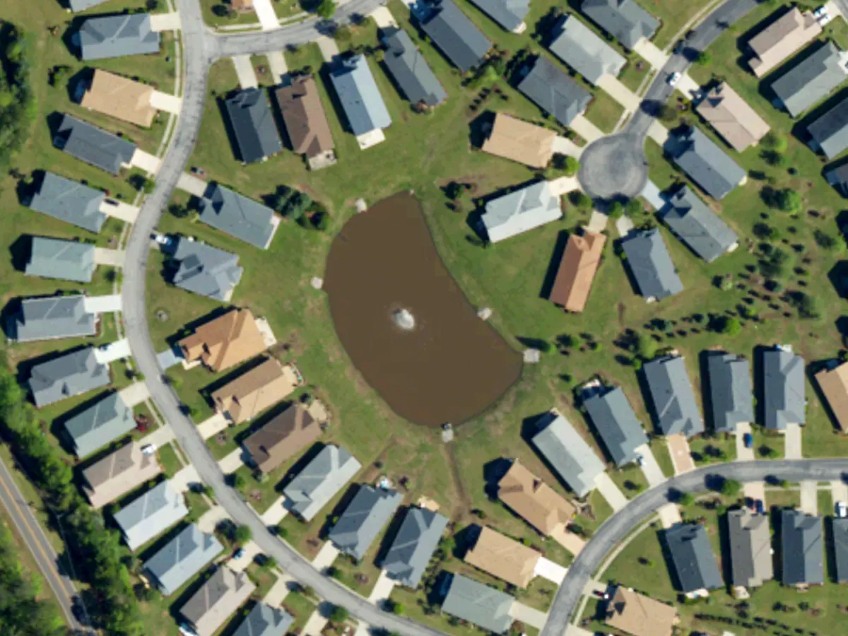 One-story homes with gray and tan roofs and wide lawns around a pond and a curving street in Seasons at Prince Creek West, seen from above