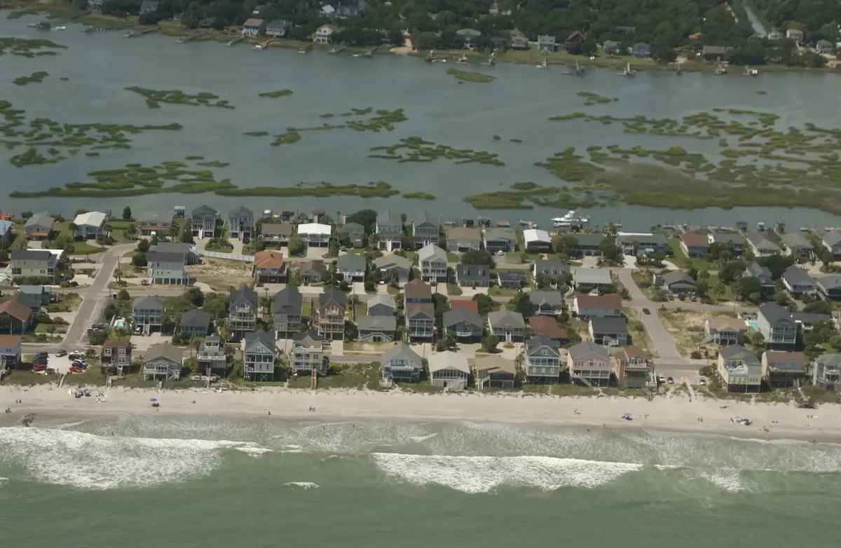 Beach houses in rows between the ocean beach and a wide marsh with tidal creeks and boat docks at Garden City Beach, seen from a plane over the ocean